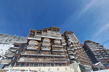 Angular, multi-story ski resort buildings with wood accents and snow-dusted balconies under a vibrant blue sky, set in a snowy mountain landscape.