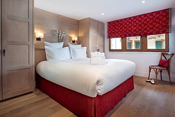A cozy bedroom with a white bed, red bed skirt, light wood paneling, built-in wardrobes, and a red patterned window shade.