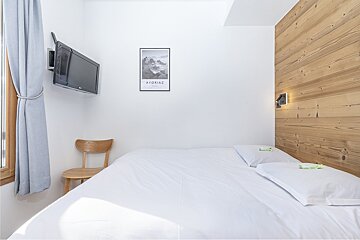 A bright bedroom with a white bed, white walls, and a wooden accent wall. A mounted TV, window with blue curtains, and an