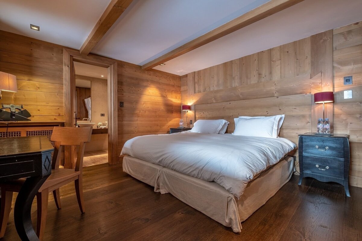 A cozy, wood-paneled bedroom featuring a large bed with white linens, side tables with lamps, a desk, and a glimpse of a bathroom.
