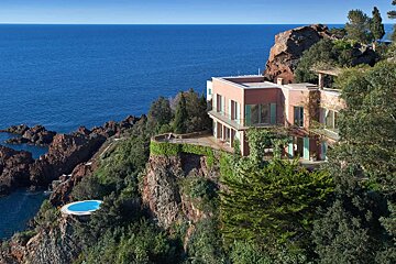 Luxurious pink villa on a green cliff, overlooking a deep blue sea. A secluded pool is built into the lower rocks.
