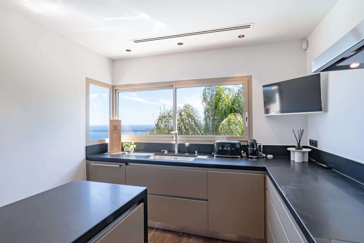 A modern kitchen with a large window offering a beautiful sea and palm tree view. It features sleek countertops, a sink, toaster, and kettle.