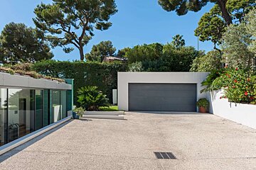 A modern building with a gray garage door, a large concrete driveway, and lush greenery, all under a bright blue sky.