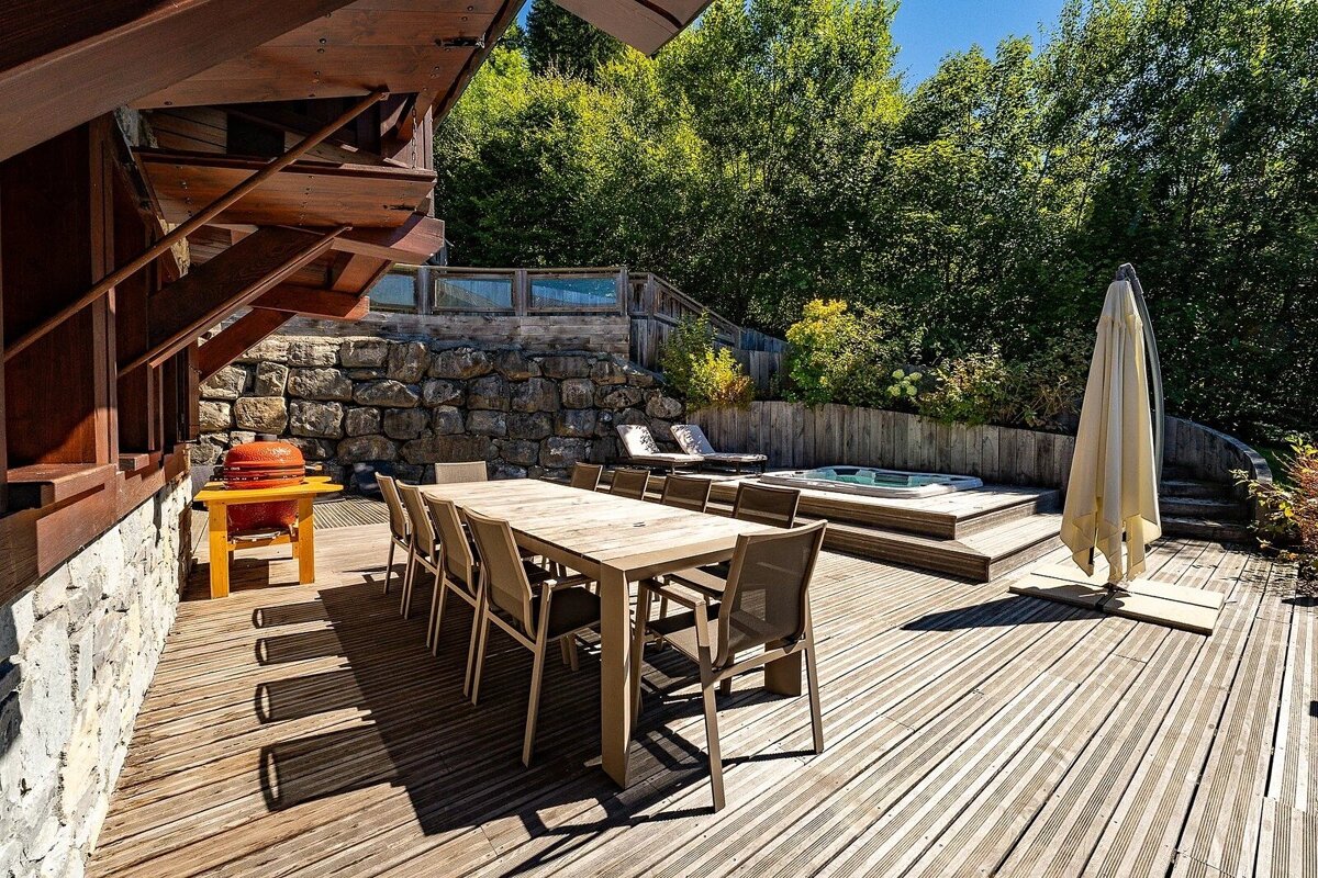 A spacious wooden deck features an outdoor dining table, red grill, and hot tub, surrounded by a stone wall and lush greenery under a clear sky.