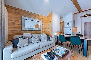 A modern open-plan living and dining room with a light-colored sofa, wooden accent wall and mirror, and a dining table with teal chairs.