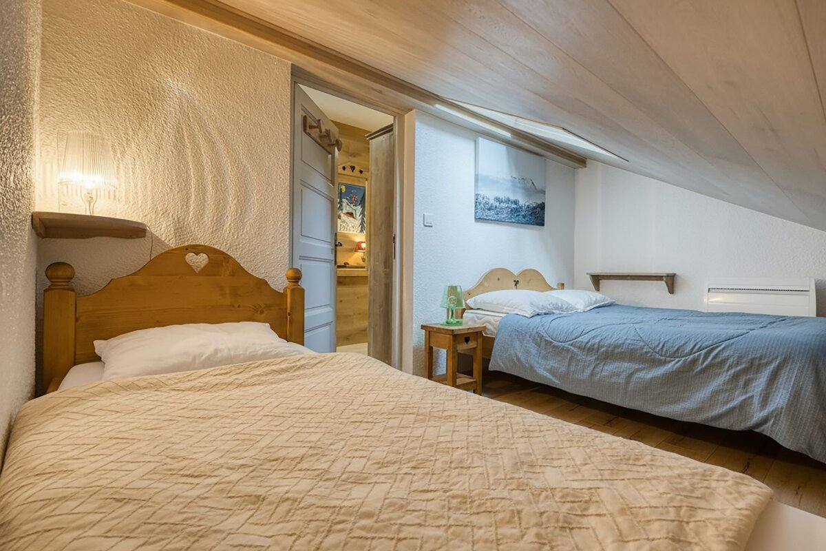 A cozy attic bedroom features two single beds with wooden headboards, a slanted wooden ceiling, and a doorway leading to another room.
