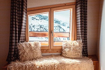 Cozy window seat with fuzzy pillows and plaid curtains, overlooking snowy mountains from a rustic cabin.