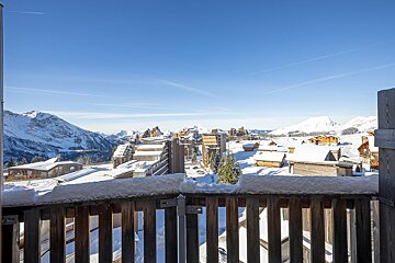 A snow-covered wooden balcony overlooks an alpine ski resort village with snowy chalets and majestic mountains under a bright blue sky.