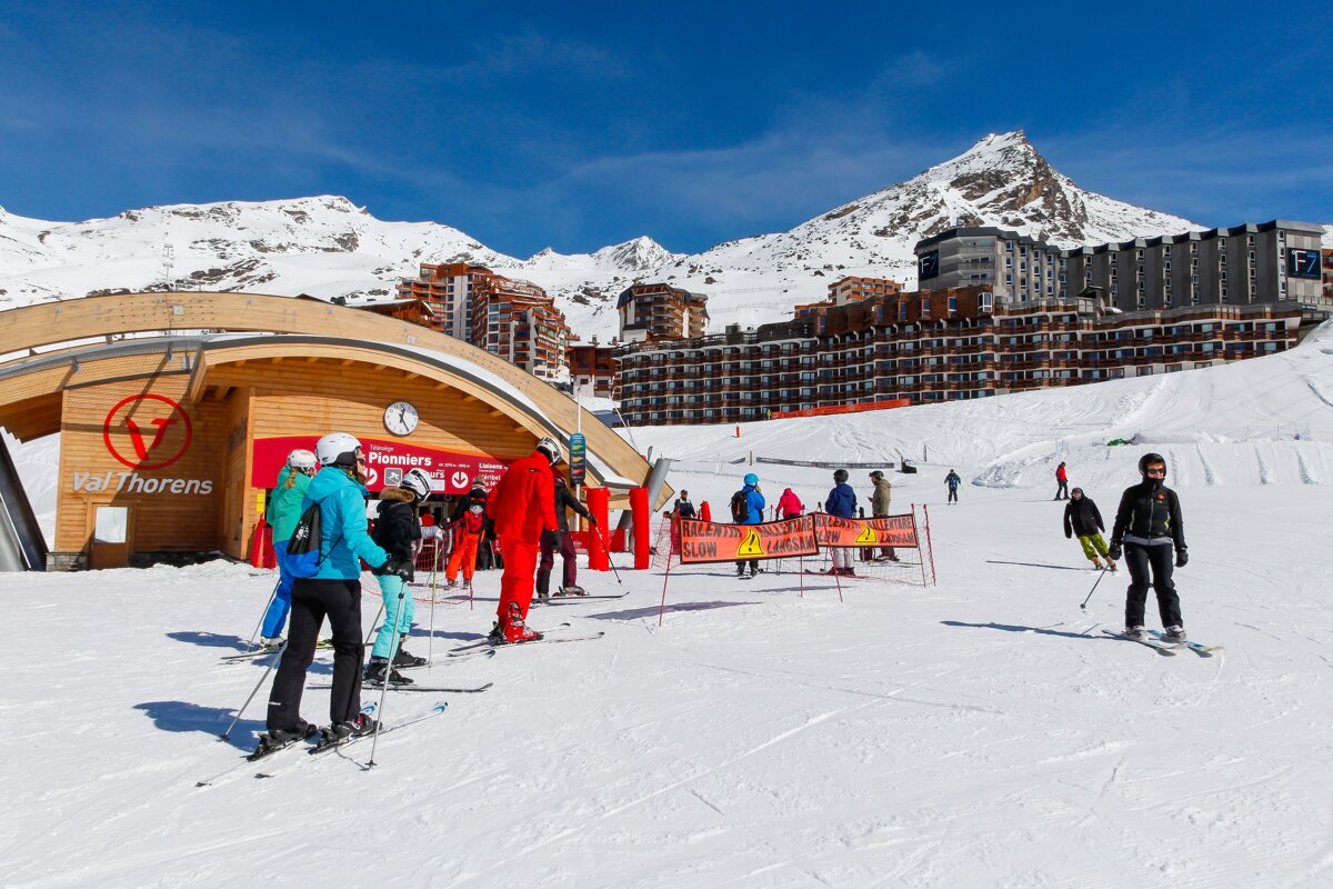 Skiers gather at the Val Thorens resort lift station on a sunny day, with snow-covered mountains and large buildings in the background.