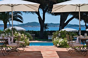 An elegant outdoor dining terrace overlooks a bright blue sea. Two white umbrellas frame tables set for dining, surrounded by lush white roses, palms, and a small pool.