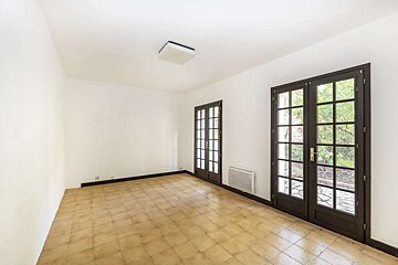 Bright, empty room with white walls, beige tiled floor, two dark French doors, a wall heater, and a ceiling light.