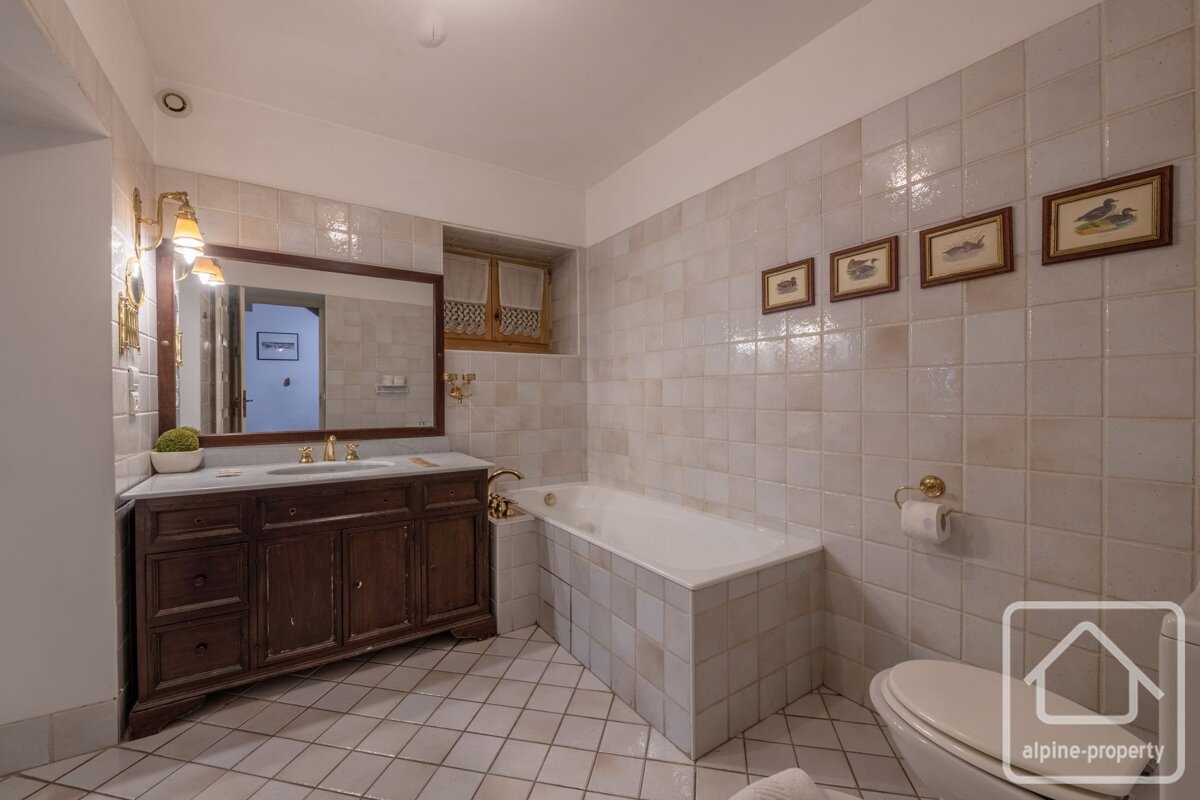 Traditional tiled bathroom with a dark wood vanity, large mirror, built-in bathtub, and toilet. Features small framed art and brass accents.