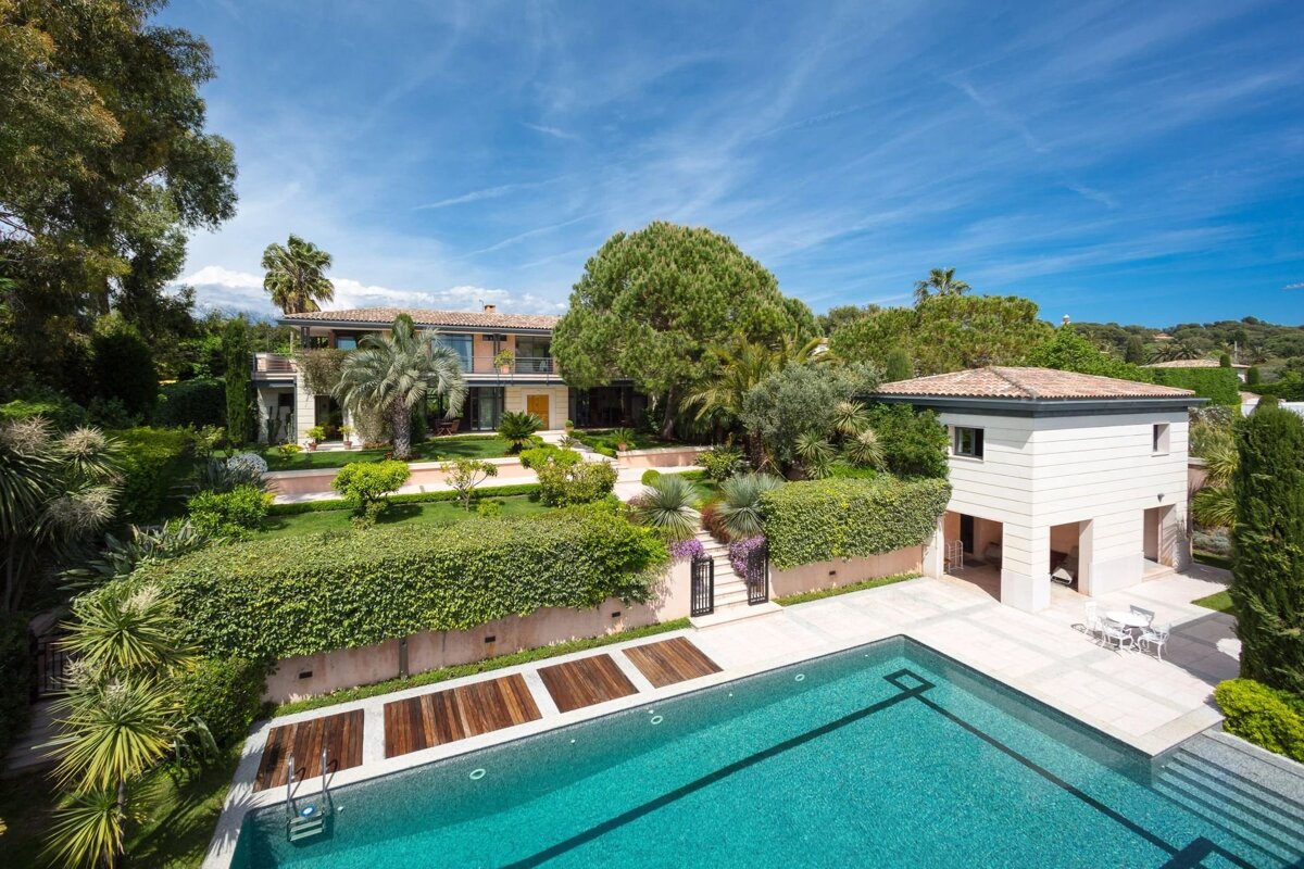 Aerial view of a luxury villa with a large swimming pool, lush green gardens, and palm trees under a bright blue sky.