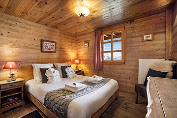 A cozy, wood-paneled chalet bedroom with a neatly made double bed, warm lighting, and a window overlooking snowy mountains.