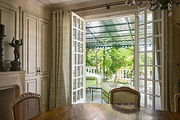 An elegant dining room with French doors opens to a serene, covered patio featuring lush greenery, outdoor seating, and natural light.