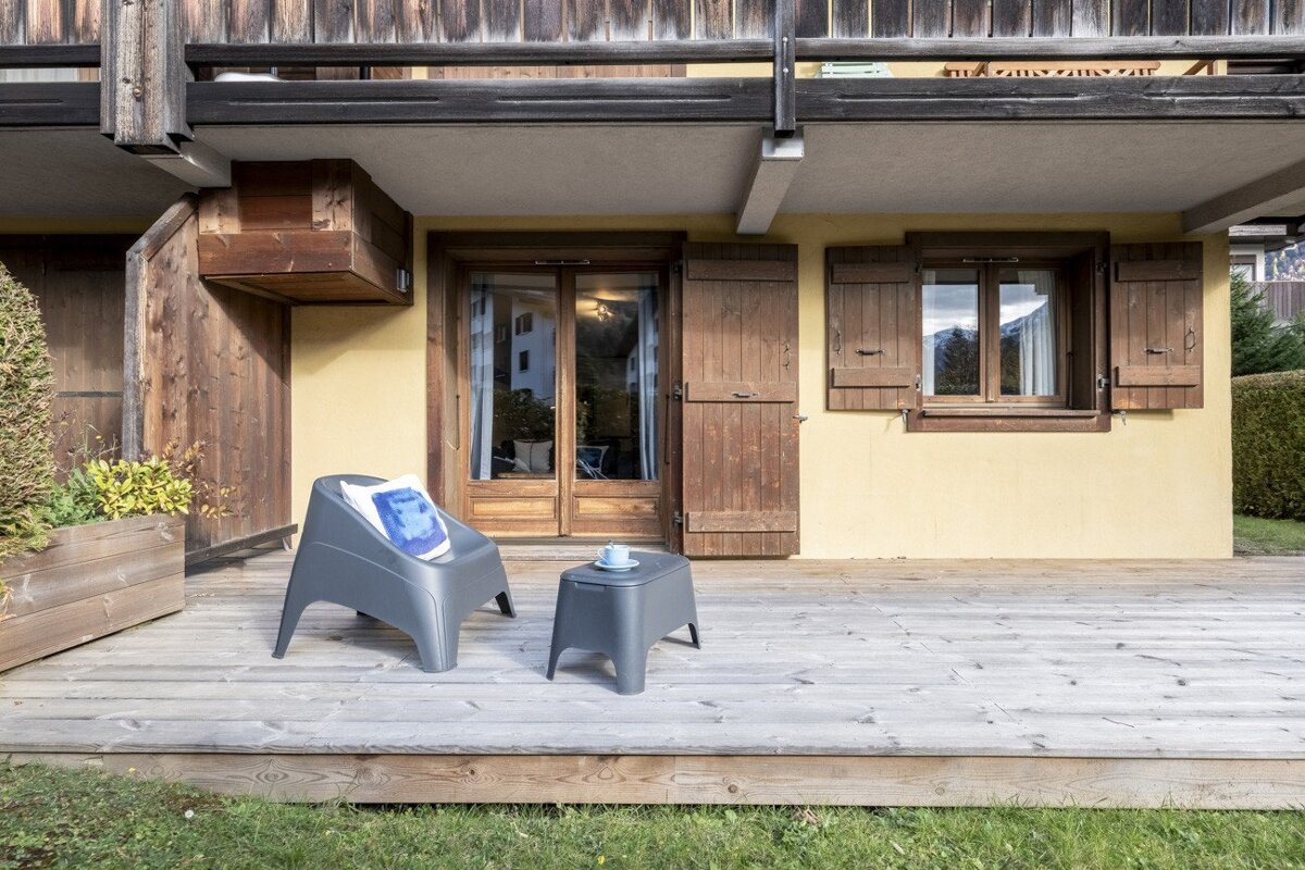 A cozy wooden deck with grey outdoor furniture outside a yellow house, featuring rustic wooden doors and window shutters, with green grass in the foreground.