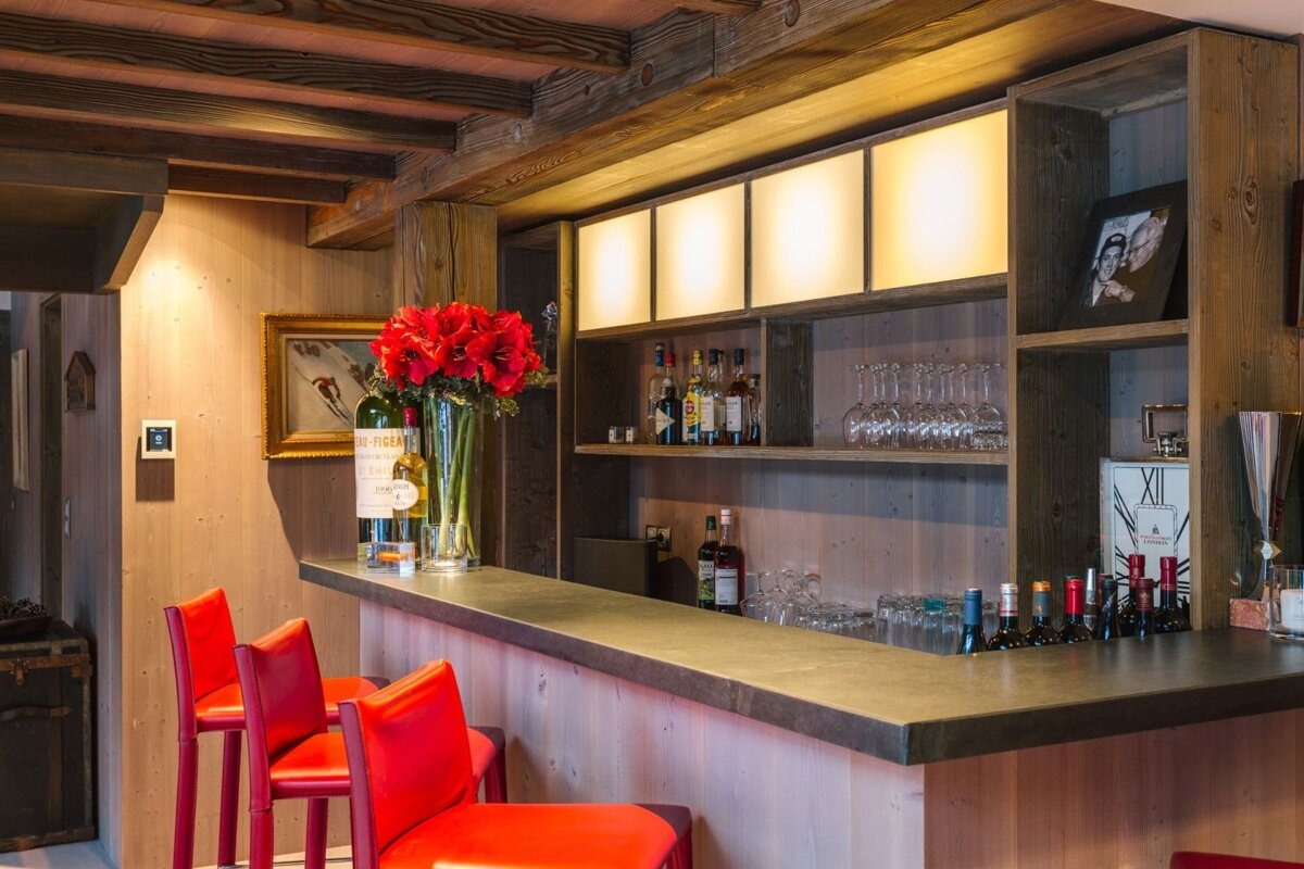 A rustic wooden home bar features red bar stools, exposed beams, and illuminated shelves stocked with liquor and glassware, accented by red flowers.