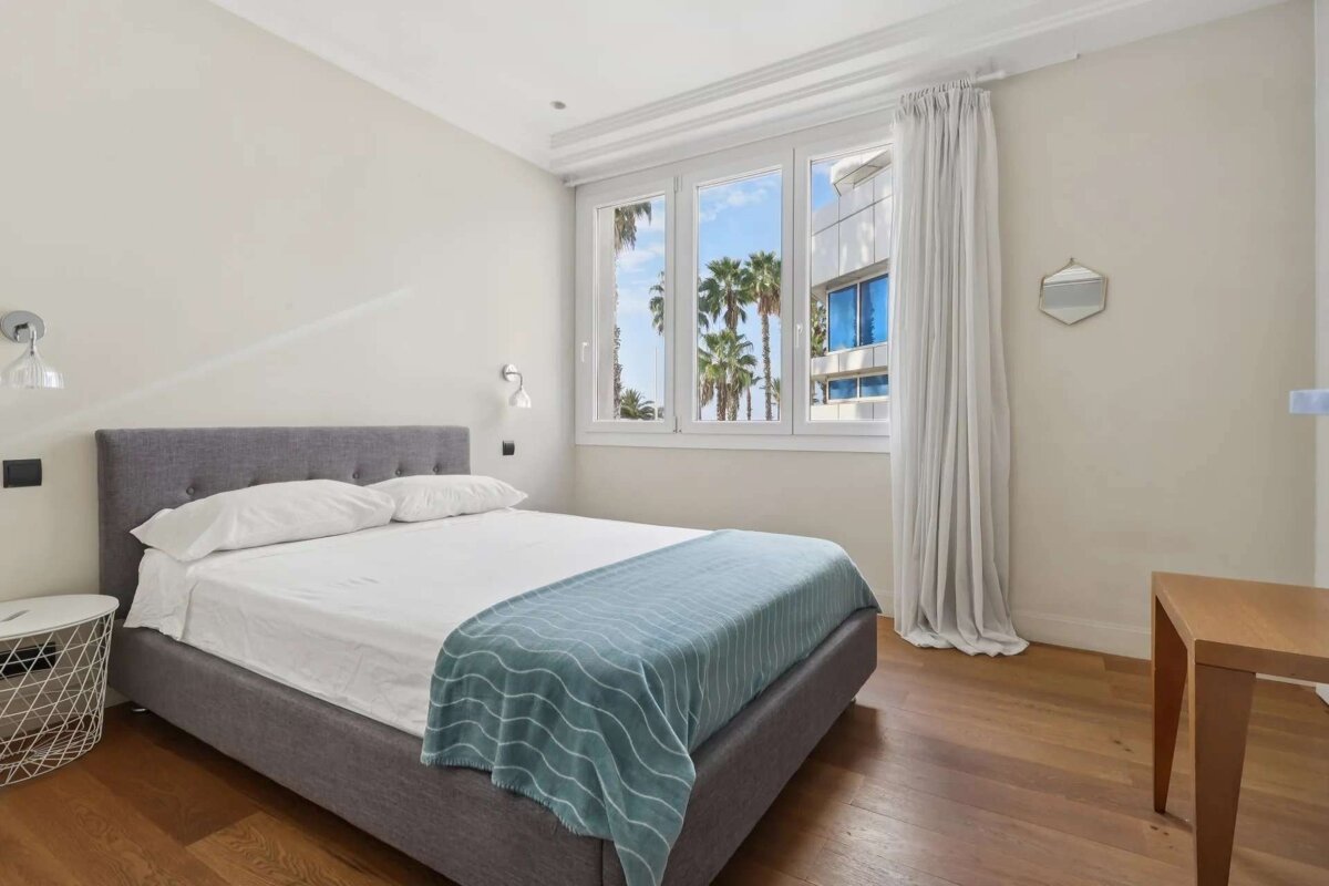 A modern bedroom with a gray bed, white bedding, blue throw, and hardwood floors. A large window overlooks palm trees, brightening the room.