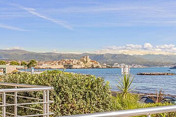 Scenic view of Antibes, France, featuring its historic fort and town beside the blue Mediterranean Sea, backed by mountains under a clear sky.