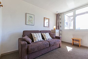 A cozy living room features a brown sofa with striped cushions, white walls, and a large window. A framed picture and mirror adorn the wall, with a small wooden stool on brown carpet.