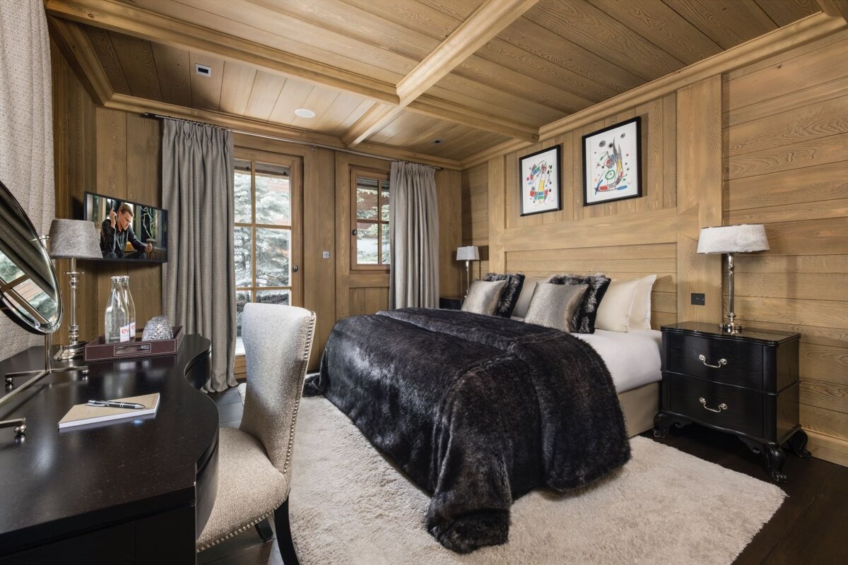 A luxurious, wood-paneled bedroom features a large bed with a dark fur throw, a desk, and windows, creating a cozy chalet ambiance.