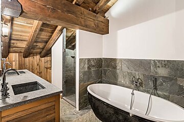 A rustic-modern bathroom with exposed wooden beams, wood paneling, and dark stone tiles. It features a double vanity, a free-standing tub, and a walk-in shower.