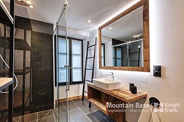 A chic bathroom featuring a glass shower, rustic wooden vanity with a vessel sink, and a large, backlit mirror, contrasting dark tiles with light walls.