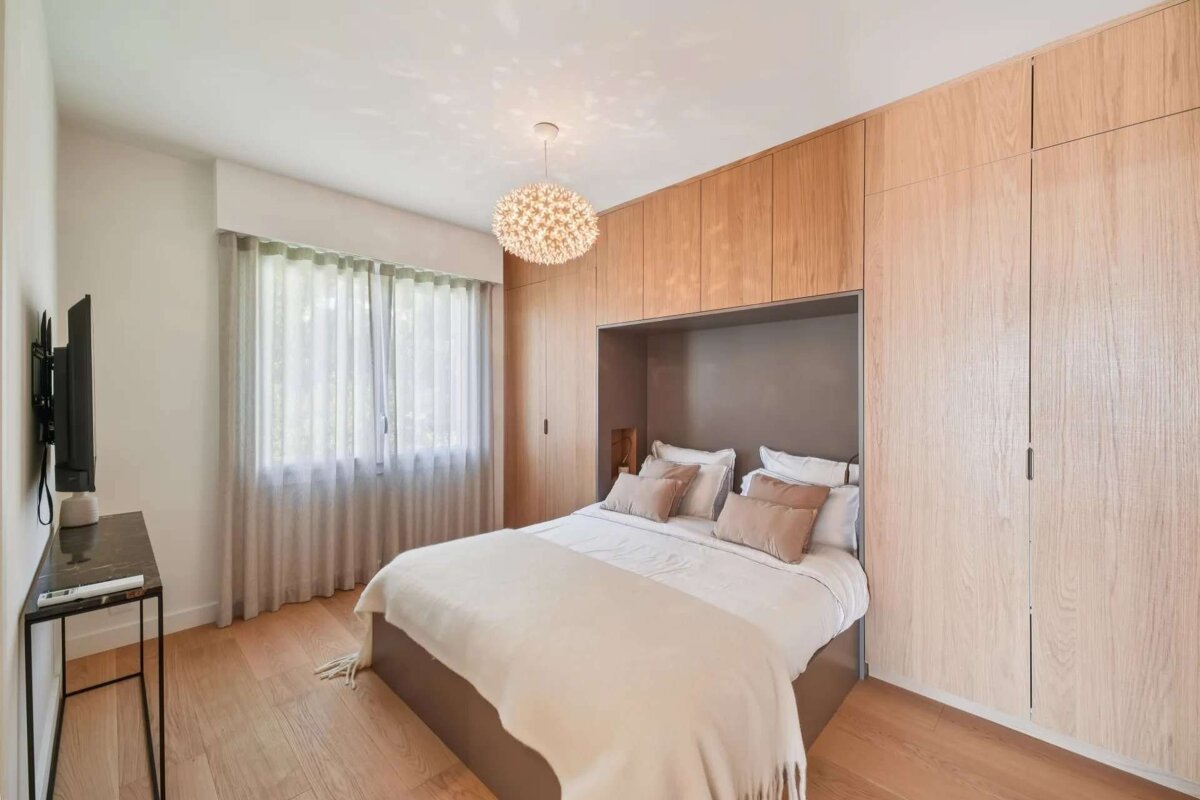 A modern bedroom features a bed framed by light wooden built-in wardrobes, a window with sheer curtains, wooden floor, and a spherical pendant light.