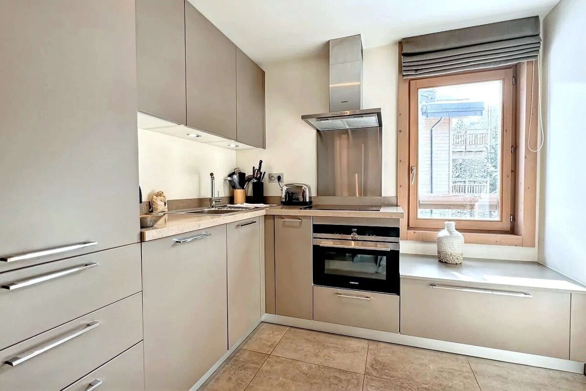 A modern L-shaped kitchen featuring sleek beige cabinets, stainless steel appliances, a large window, and tiled flooring.
