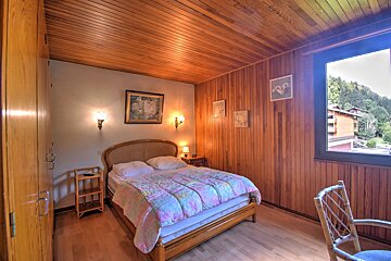 A rustic bedroom with extensive wood paneling on walls/ceiling, a double bed with a colorful quilt, wood floor, and a window offering a mountain view.