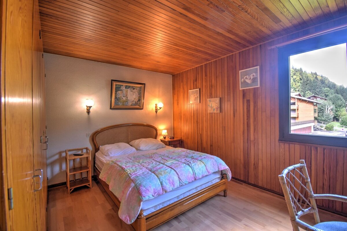 A rustic bedroom with extensive wood paneling on walls/ceiling, a double bed with a colorful quilt, wood floor, and a window offering a mountain view.