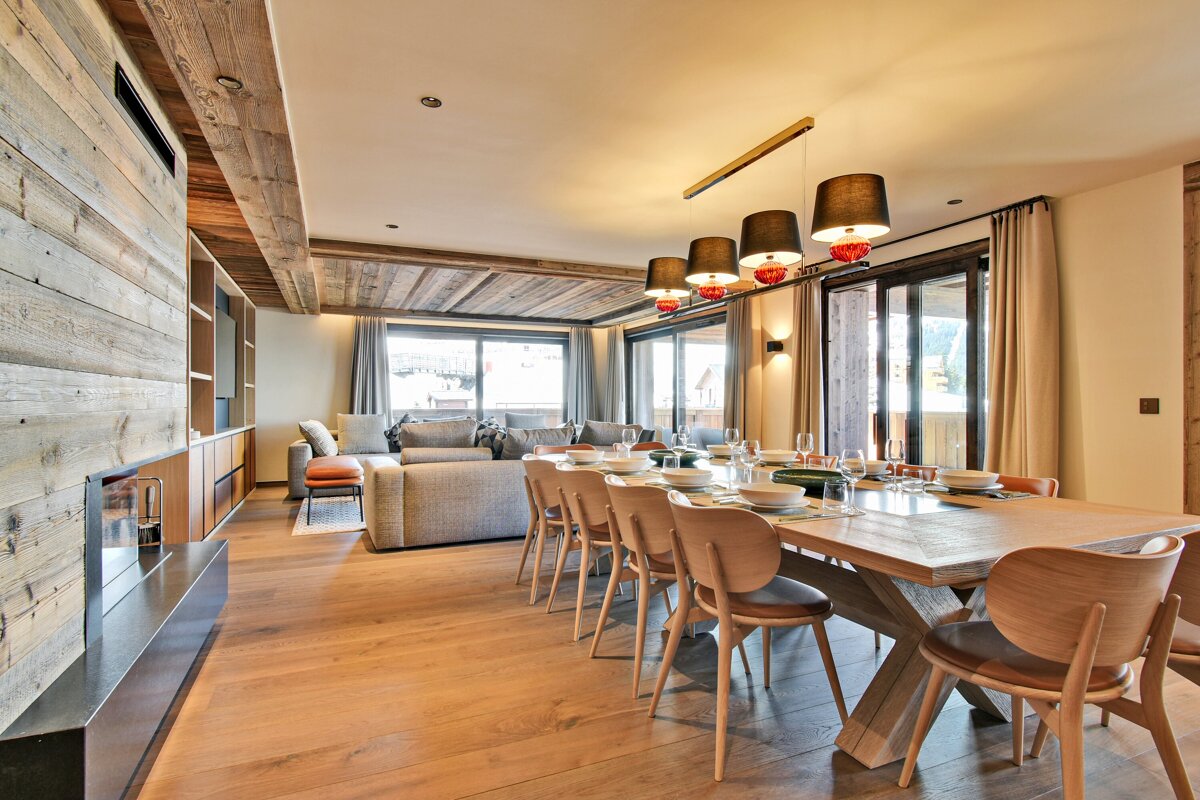 Modern rustic chalet interior with a large dining table, comfortable living room, exposed wooden beams & paneling, and big windows.