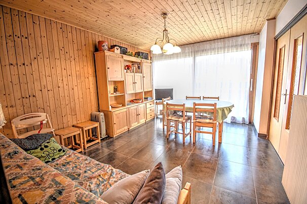 A warm, rustic living/dining room with wood-paneled walls and ceiling, a sofa, large wooden cabinet, and dining table. Bright window and tiled floor.