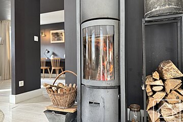 A modern wood-burning stove with visible flames and stacked firewood creates a cozy ambiance. A dark-walled room with wooden furniture is visible in the background.