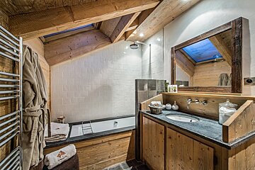 Cozy, rustic bathroom with exposed wood beams, a white-tiled bath/shower area, wooden vanity, sink, and a skylight.
