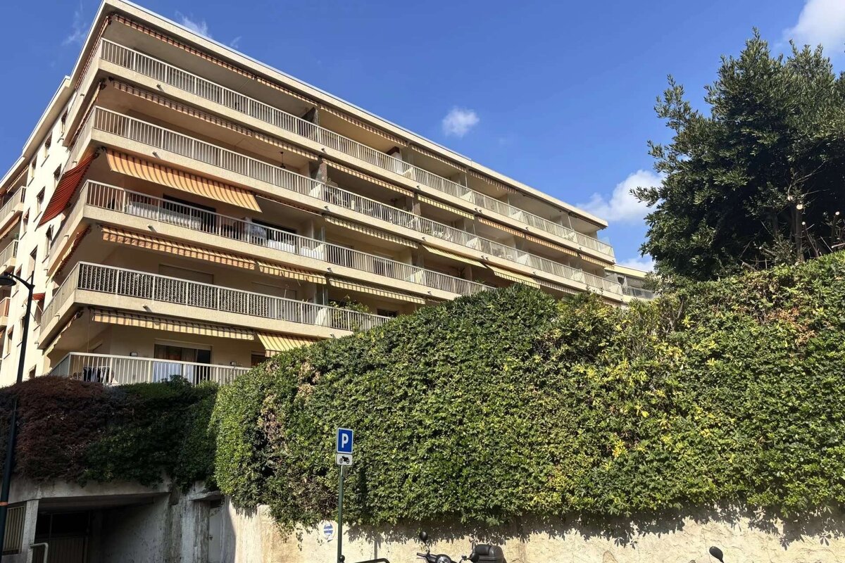 A multi-story apartment building with balconies & striped awnings, fronted by a lush green hedge and a parking sign, under a blue sky.