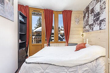 A cozy, wood-paneled bedroom with a white bed, red curtains, and a bay window showing a snowy village. Deer decor and a cowhide patterned wall add to the chalet feel.