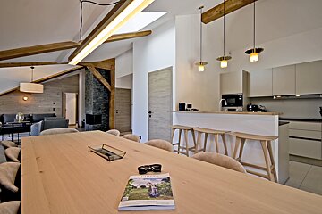 Modern open-plan chalet with a long wooden dining table, kitchen island with stools, and vaulted ceiling with wooden beams.