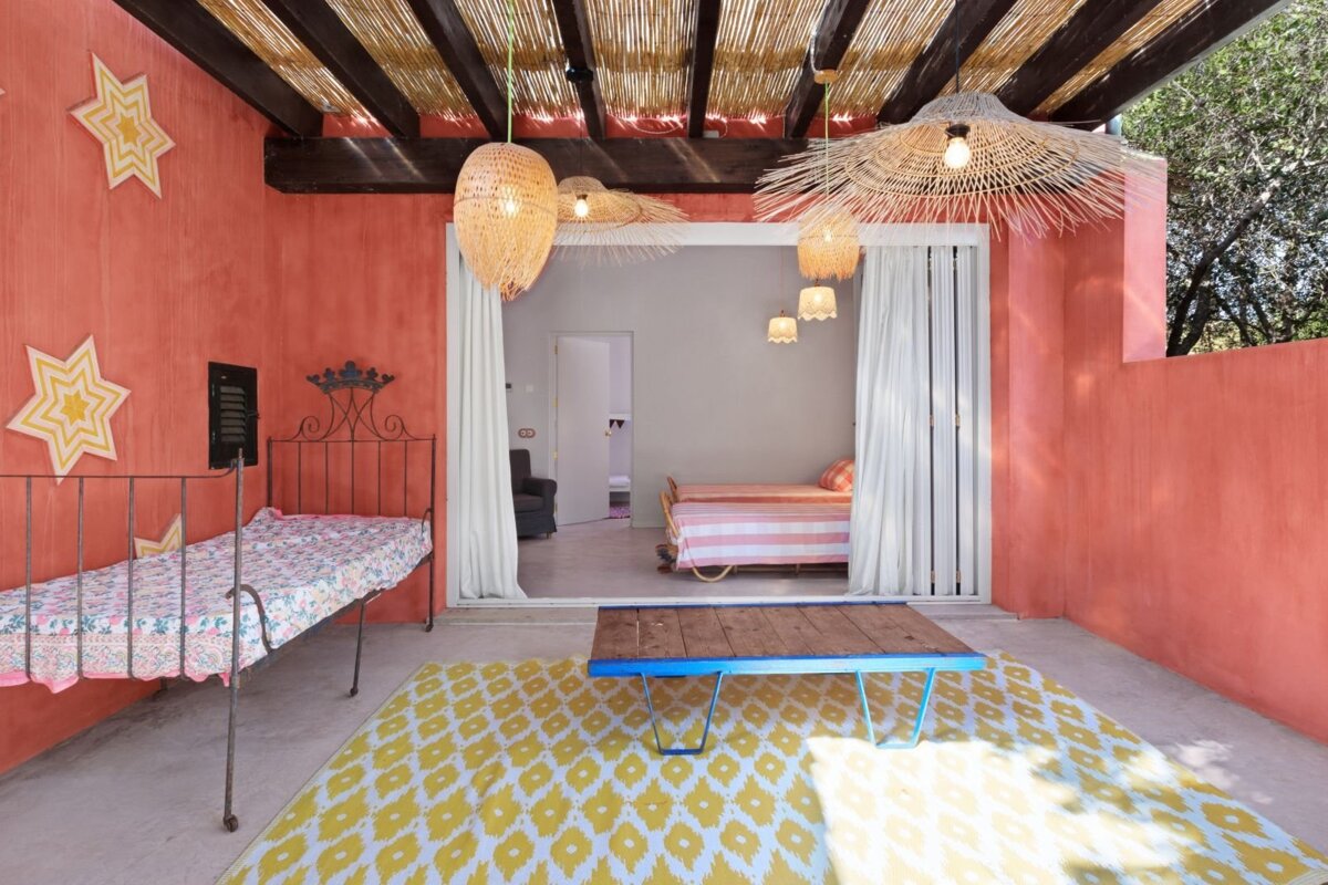 A vibrant coral room with a bamboo ceiling, woven lamps, and two daybeds. A yellow rug and open doorway create a rustic, airy space.