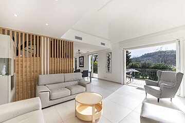 Bright, modern living room with white floors/walls, light grey sofas, wooden partition, and a dining area, opening to a balcony with mountain views.