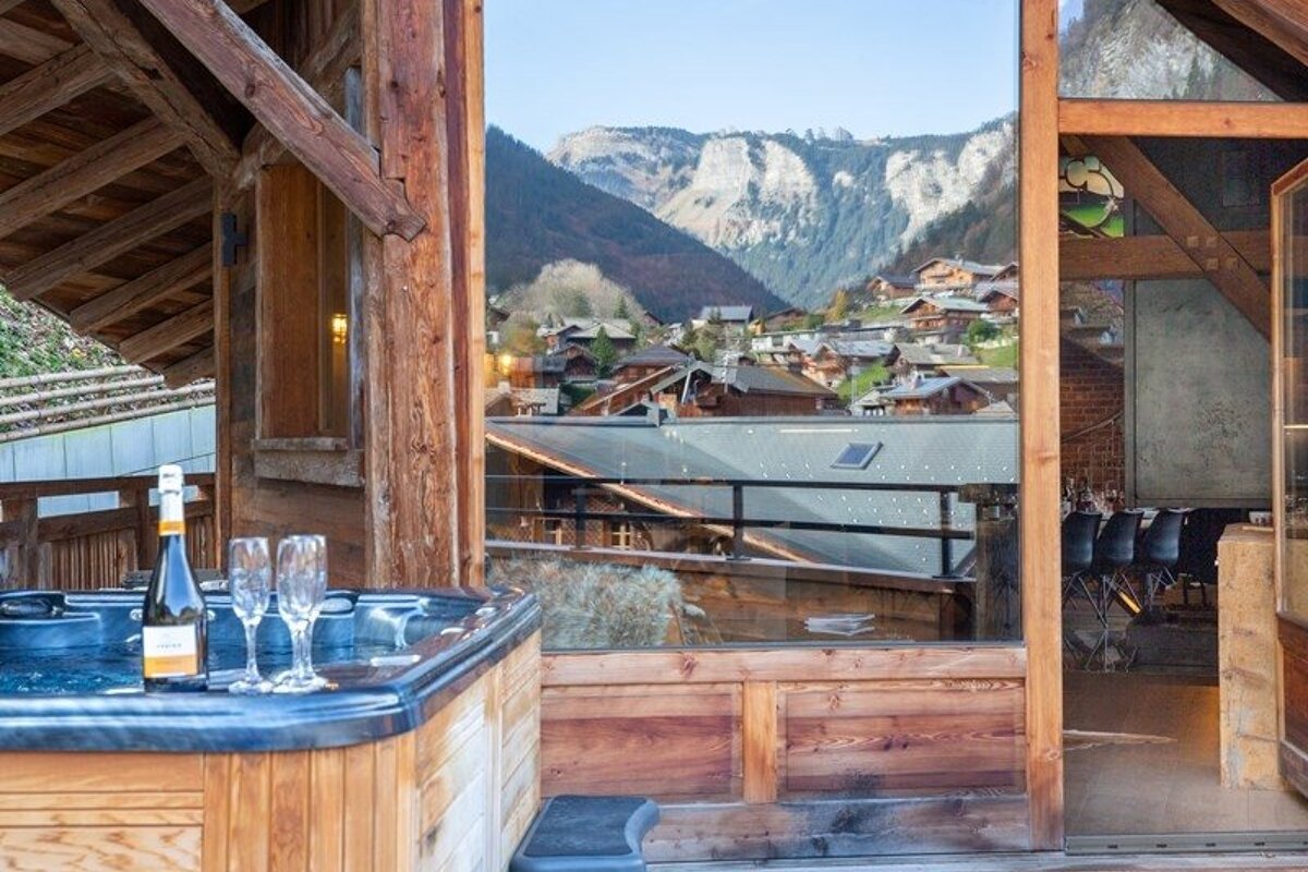 Hot tub with champagne on a chalet balcony overlooking an alpine village and mountains.