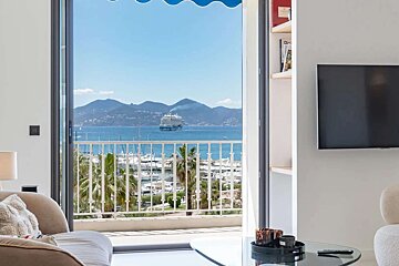 A vibrant apartment balcony view overlooks a blue sea, distant mountains, a cruise ship, and a bustling marina with yachts and palm trees.