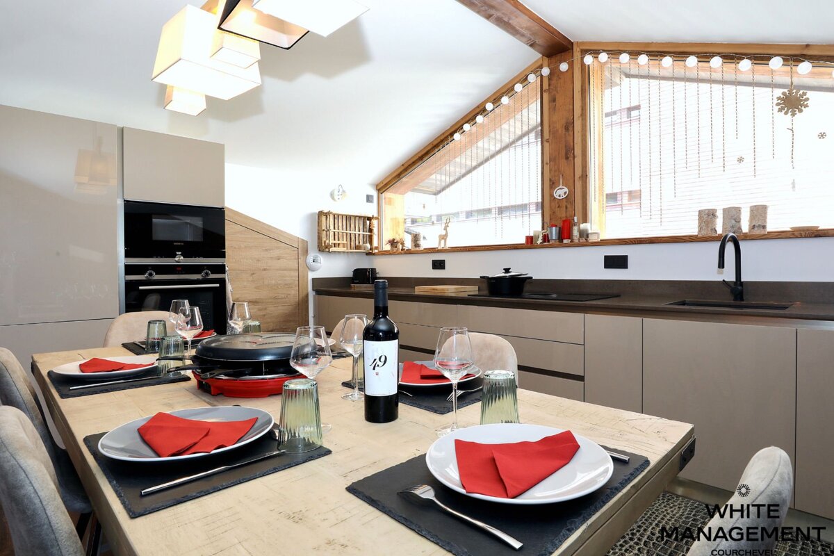 A modern kitchen and dining area with a table set for a raclette meal, wine, and red napkins. Features neutral tones, wood accents, and a festive window.