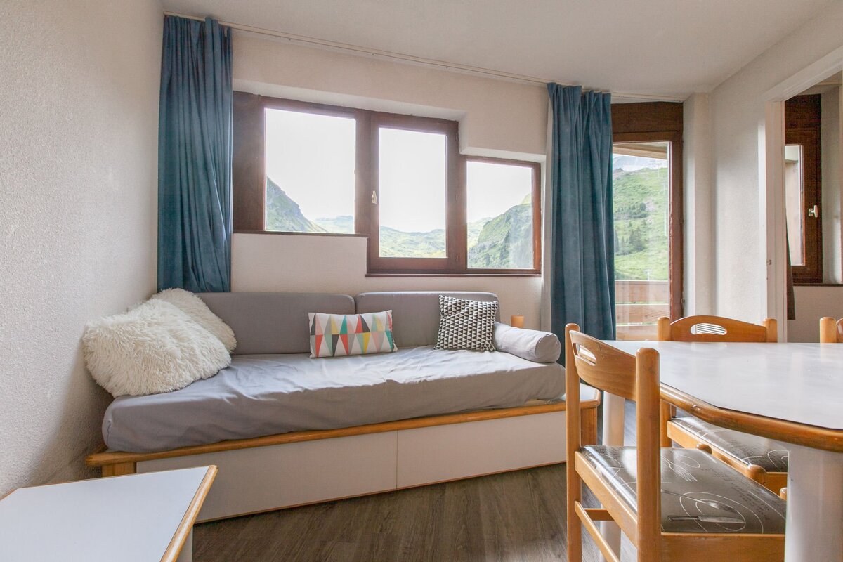 A room with a grey daybed, dining table, and large windows offering a panoramic view of lush green mountains. Blue curtains frame the scene.