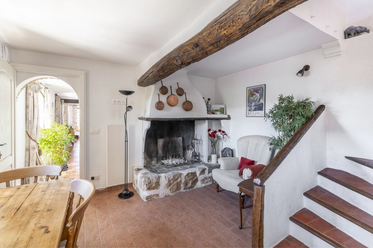 Cozy rustic room featuring a large stone fireplace, exposed wooden beam, terracotta floor, staircase, and an archway leading to another room.