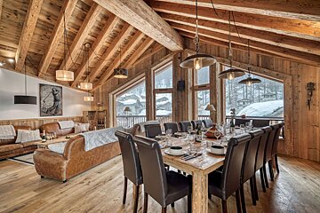 Rustic wooden chalet interior with a large dining table, leather chairs and sofas, and snowy alpine views through big windows.
