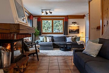 A warm, chalet-style living room with a glowing fireplace, large gray sectional, and window overlooking trees. Rustic wood accents and cozy cushions complete the scene.