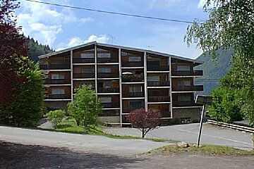 A multi-story brown and white building with balconies sits amidst green trees and mountains under a cloudy sky. A paved road is in the foreground.