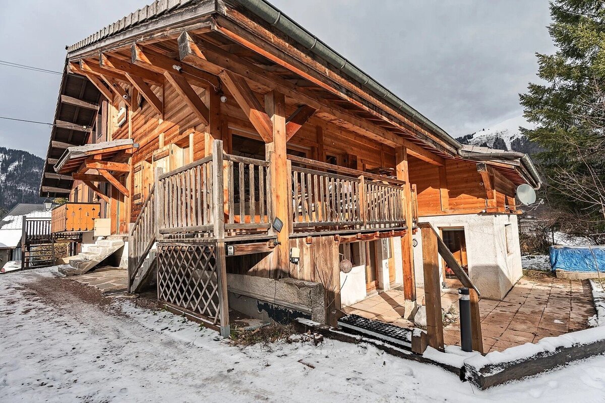 A rustic wooden chalet with exposed beams and a balcony, surrounded by snow, with snowy mountains in the background under a cloudy sky.