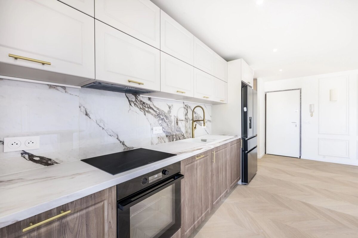 Modern kitchen with white and wooden cabinets, marble countertops, gold hardware, and herringbone wood floors.